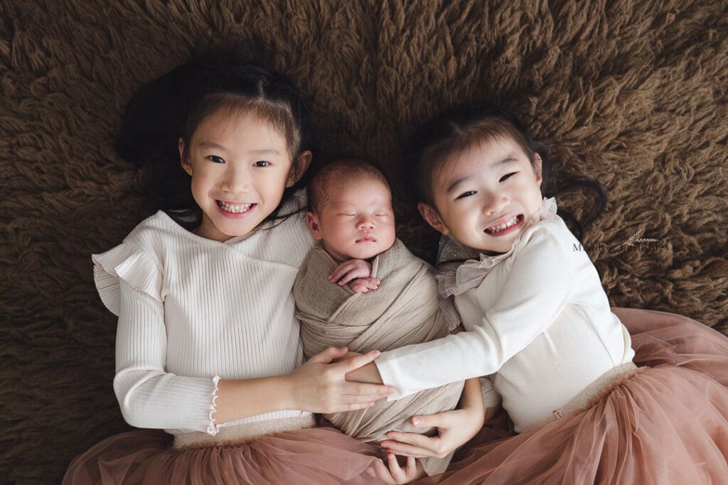 Newborn photoshoot elder sisters posing with newborn baby boy on brown flokati fur rug