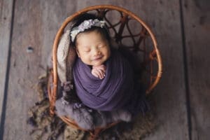 Newborn photography baby wrapped in purple posing on a vintage rattan chair prop