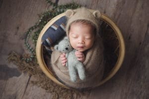 newborn baby with bear hat holding large grey bear soft toy in vintage round rattan stool